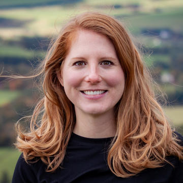 A red-haired white woman stands smiling in the photo, with a view of a valley behind her